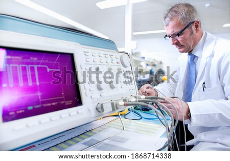 An Engineer in lab coat and eyeglasses under selective working at an electrical test bench next to an oscilloscope