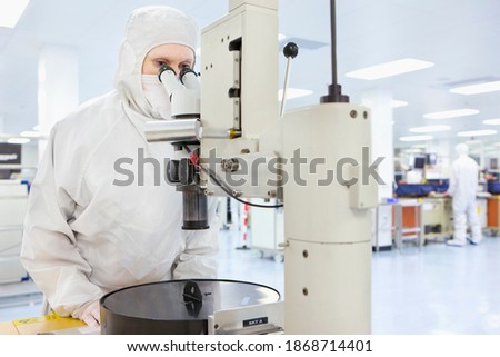 A scientist in a clean suit using an industrial microscope in the silicon wafer manufacturing laboratory