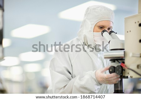 A horizontal scientist in a clean suit adjusting the lens while carefully examining the silicon wafer under a microscope in a laboratory