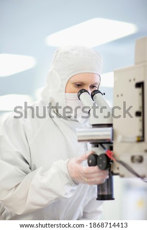 A vertical scientist in a clean suit adjusting the lens while carefully examining the silicon wafer under a microscope in a laboratory