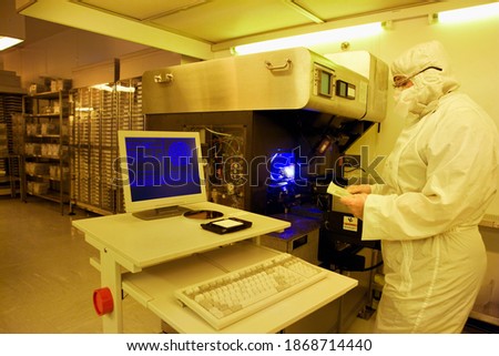 An engineer in a clean suit reading paperwork while examining a silicon wafer near the computer system in a special laboratory with warm light