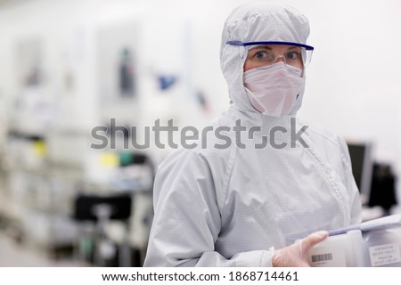 A horizontal portrait of a scientist in a clean suit carrying a container in the silicon wafer manufacturing laboratory