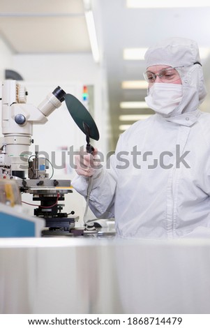 A Scientist in a clean suit examining the silicon wafer with the help of a vacuum handling tool near the microscope in a special laboratory