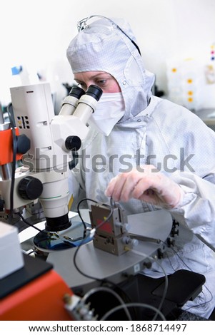 A vertical view of a scientist in clean suit carefully examining the silicon wafer while making adjustments under a microscope in a special laboratory