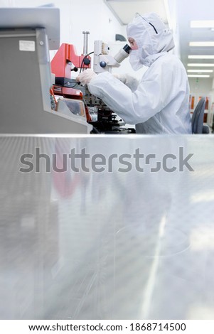 A distant vertical view of a Scientist in clean suit examining the silicon wafer under a microscope in a special laboratory