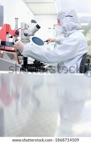 A vertical view of a scientist in clean suit examining the silicon wafer while sitting next to a microscope in a special laboratory