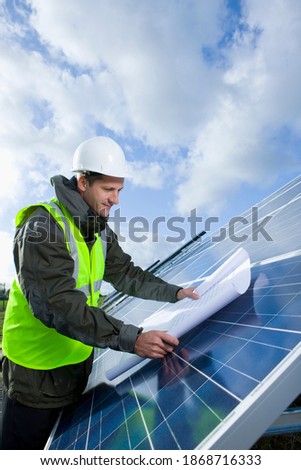 A vertical shot of an engineer looking at blueprint on a large solar panel with sky in background.