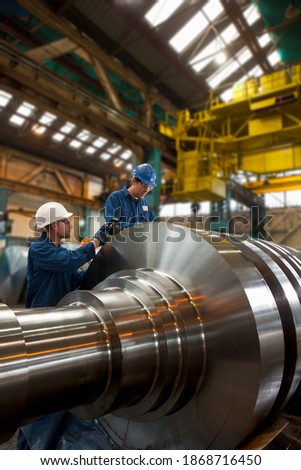 A vertical shot of two engineers in coverall cleaning a big metal machinery in a warehouse.