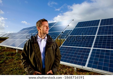 A medium shot of a happy farmer looking sideways while standing in front of big solar panels.