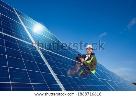 A low angle medium shot of an engineer standing next to a big solar panel and looking sideways while holding a tablet.