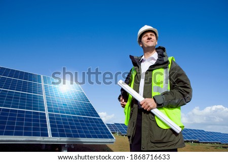 A low angle medium shot of a happy engineer with blueprint looking up while standing in front of large solar panels.