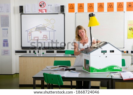A wide shot of a female teacher adjusting solar panels on a model house in a science class.