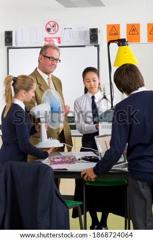 A vertical shot of a teacher watching young students in school uniforms working with wind turbine model in a science class.