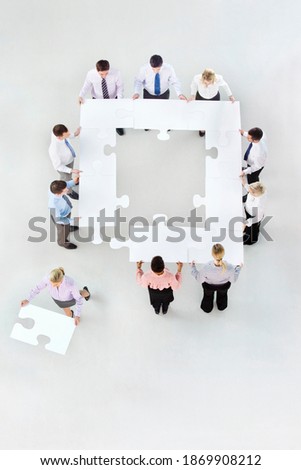 Overhead shot of businesspeople forming a square pattern with large jigsaw puzzle pieces with a businesswoman walking away with a piece.
