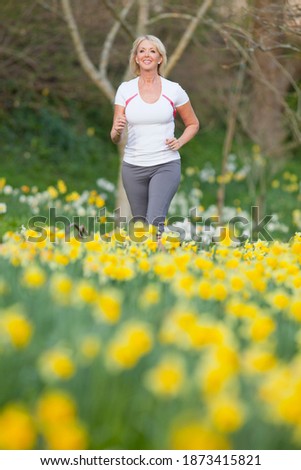 Vertical three quarter length low angle shot of a senior woman running by a daffodil field smiles at the camera on an overcast day .