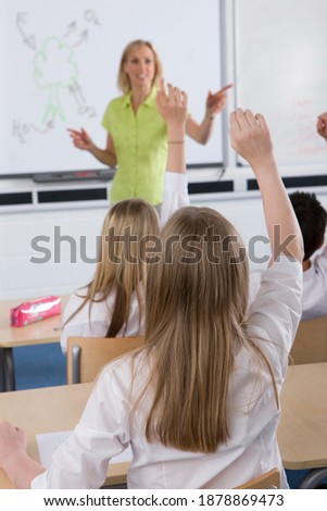 A vertical rear view of school girls raising their hands in the air to answer a question posed by their teacher in selective focus