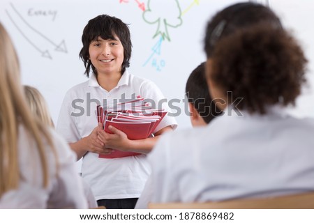 Front view of a�smiling�Student holding test booklets before passing out to his classmates�in a background of a white board�