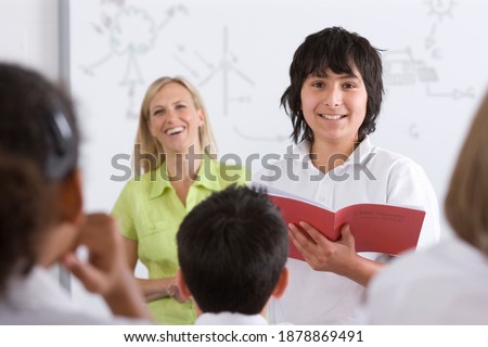 Front view of a school boy in selective focus reading out of test booklet to classmates sitting in the foreground and the�teacher standing in the background�