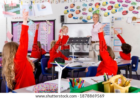 Science teacher and school girl holding a solar panel while other students raising their hands in the air to answer a question asked to the entire class