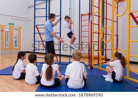 Gym teacher helping a school boy in sports uniform to climb a rope in a school gymnasium while other students sitting down and watching
