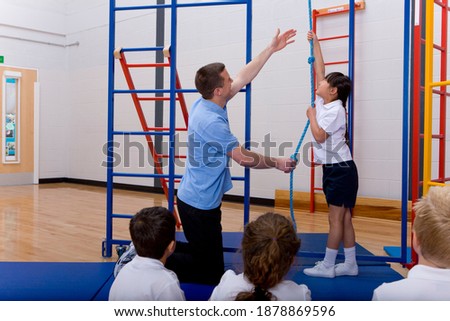 Gym teacher helping a school girl in sports uniform to climb a rope while other students sitting down and watching in the foreground