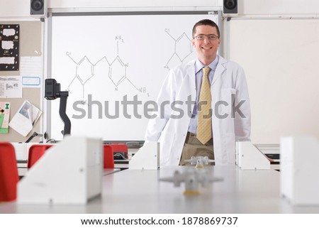 A chemistry teacher in a school laboratory wearing a Lab coat standing in front of a white board with a chemical formula written over it.