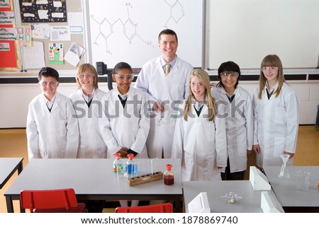 high angle view of a proud Chemistry teacher in a school lab smiling at the camera while standing with a group of smart students wearing lab coats and safety goggles