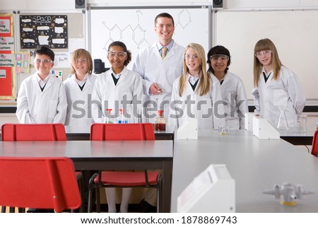 A proud Chemistry teacher in a school lab smiling at the camera while standing with a group of smart students wearing lab coats and safety goggles