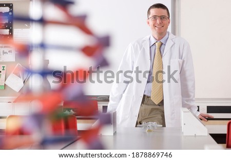 A medium wide shot of a chemistry teacher smiling at the camera in a school lab wearing a Lab coat with a DNA model placed in the foreground