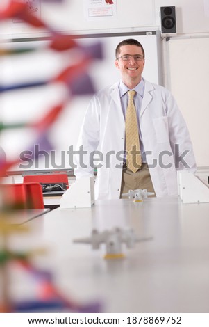 A vertical front view of a chemistry teacher smiling at the camera in a school lab wearing a Lab coat with a DNA model in the foreground