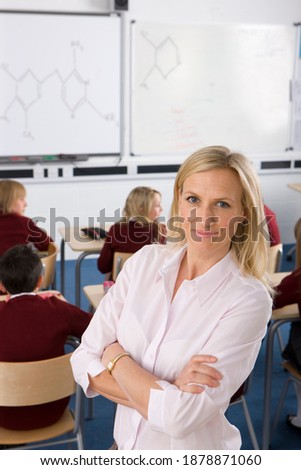A high angle vertical shot of a Proud teacher smiling and posing with her arms crossed on a blurred background of a white board and students studying on desk