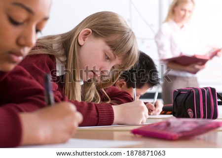 Sideview of students sitting in a row with selective focus taking a test in an exam hall with a supervisor standing in the background