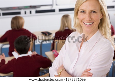 A medium high angle shot of a Proud teacher smiling and posing with her arms crossed on a blurred background of students studying on desk