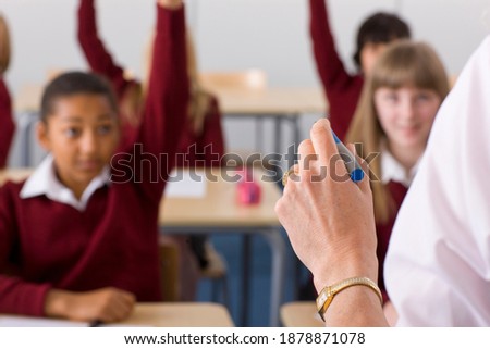 School children raising their hands in the air to answer a question posed by their teacher in selective focus