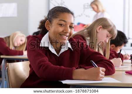 A confident girl in school uniform under selective focus taking a test on a softly blurred background of an exam hall with other students