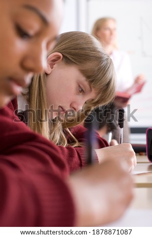 A vertical sideview of students sitting in a row with selective focus taking a test in an exam hall with a supervisor standing in the background