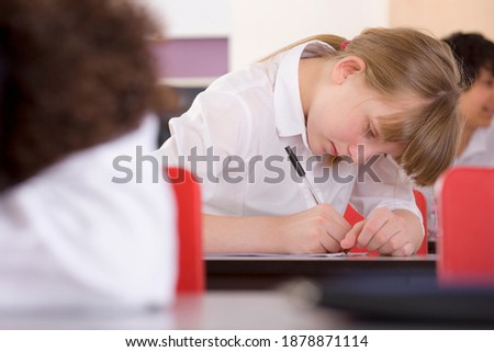 A close shot of a school girl looking down and leaning on the desk while writing on a sheet of paper in a classroom