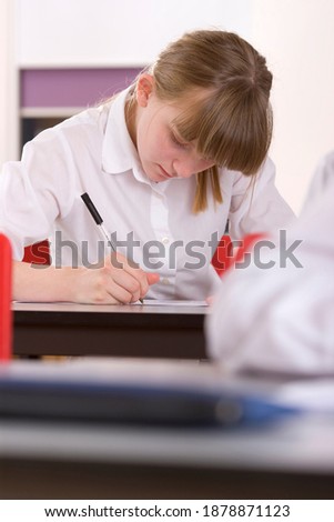 A vertical front view of a school girl looking down at her desk and writing on a sheet of paper in a classroom
