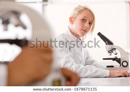 A smart school girl in a lab coat working on a microscope in a science laboratory while looking at the camera