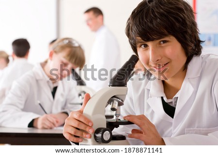A smart school boy in a lab coat working on a microscope in a science laboratory while smiling at the camera
