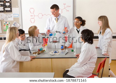 A wide angle view of a science laboratory with students in lab coats and safety goggles as they are watching an experiment conducted by the chemistry teacher