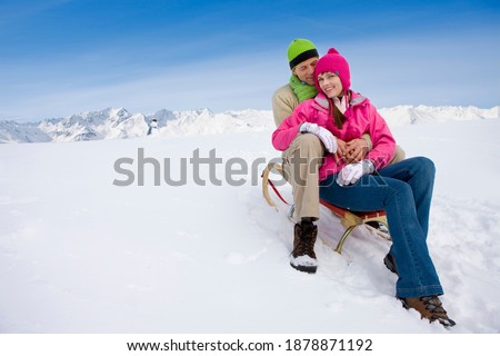 A romantic couple in winter wear sitting on a sled close to each other at the top of a ski slope