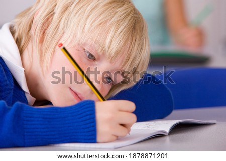 A closeup of a school boy under selective focus with blonde hair in a classroom leaning on the desk while looking down and writing in his workbook under selective focus