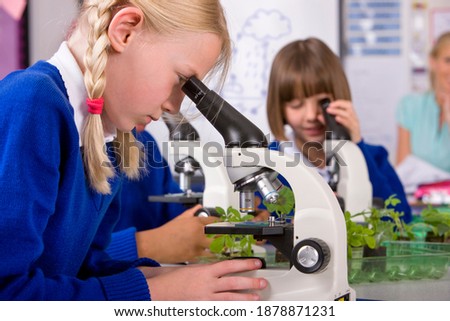 A sideview of a school girl with blonde hair in a laboratory looking into a microscope with her classmates and Teacher sitting in background