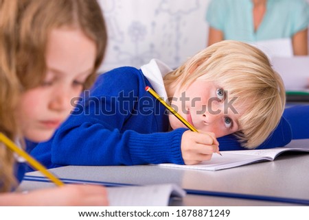 A closeup of a school boy with blonde hair in a classroom under selective focus leaning on his desk while thinking and writing answers in the workbook.