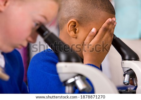 A sideview of preteen boy in a school laboratory covering his eye with one hand trying to focus his vision for looking into a microscope more clearly with another student sitting in the foreground