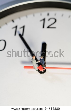Two miniature businessmen toy models shake hands to seal a business deal while balanced on the hands of a clock, overhead view