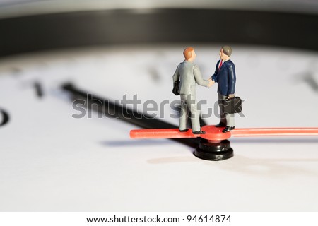 Two miniature businessmen toy models shake hands to seal a business deal while balanced on the hands of a clock, macro side view