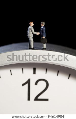 Two tiny model figures of businessmen shaking hands on top of a clock showing the time as five minutes to twelve, vertical.