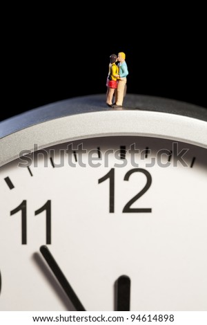 Miniature toy figurines of a young couple in a romantic embrace are balanced on the top of a clock showing five minutes to midnight.